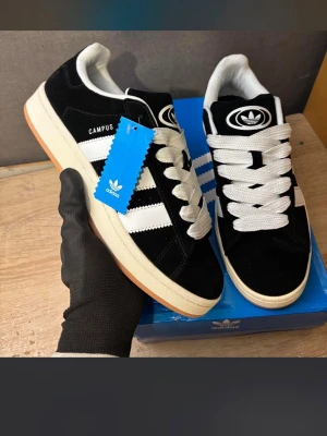 Adidas Campus svartvita sneakers - Snygga Adidas Campus sneakers i svart mocka med vita detaljer och klassiska tre ränder på sidan. Skorna har breda vita skosnören, vit sula och Adidas logga på plösen. Perfekta för dig som gillar retrostil och streetwear.