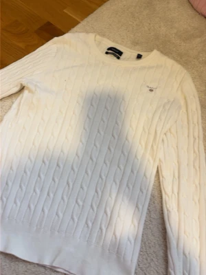 Vit kabelstickad tröja från Gant - Säljer en klassisk vit kabelstickad tröja från Gant i stretchig bomull. Tröjan har rund halsringning, långa ärmar och Gant-logga broderad på bröstet. Perfekt för dig som gillar stilrena och tidlösa plagg.