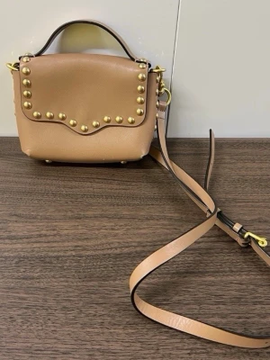 Rebecca Minkofff Beige handväska med guldfärgade nitar - Snygg beige handväska i slätt läder med guldfärgade nitar längs locket. Väskan har både ett handtag och en lång justerbar axelrem. Perfekt för dig som gillar detaljer och vill ha en trendig accessoar till din outfit.