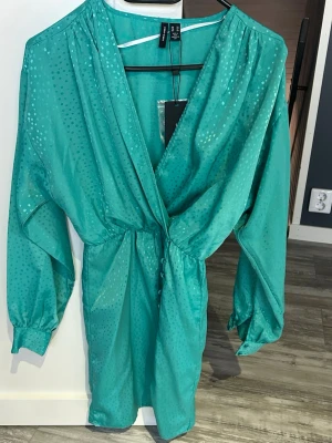 Turkos omlottklänning från Veto Moda - Snygg turkos omlottklänning från Veeo Moda med subtilt prickigt mönster och v-ringning. Klänningen har långa ärmar med breda muddar och markerad midja. Perfekt för dig som vill sticka ut med färg och stil.