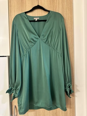 Grön omlottklänning i satin - Snygg grön kortklänning i satin med v-ringning och långa ärmar. Ärmarna har knyt vid handlederna för extra detalj. Perfekt för dig som gillar en elegant och trendig look.