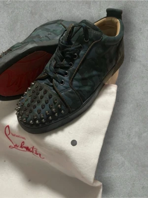 Christian Louboutin camo sneakers - Unika sneakers från Christian Louboutin i mörkgrönt camomönster med svarta nitar på tån och klassisk röd sula. Skorna har svarta snören och detaljer i svart läder. Perfekt för dig som vill sticka ut med edgy design och exklusiv känsla. #loubotin #valentino #lanvin #skor