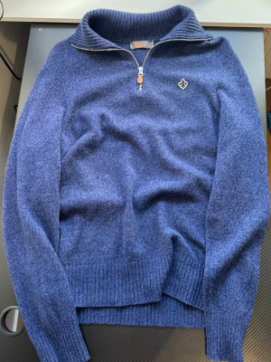 Blå stickad tröja från Morris - Snygg blå stickad tröja från Morris med halvzip och hög krage. Tröjan har ribbade muddar, diskret Morris-logga på bröstet och är gjord i mjukt material som känns lyxigt. Perfekt för lager på lager och chill stil.