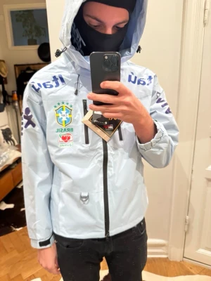 Nike Brasil vindjacka med huva - Ljusblå vindjacka från Nike med Brasil-logga och sponsormärken som Itaú och Vivo. Jackan har huva med dragsko, två bröstfickor med dragkedja och justerbara ärmslut. Tillverkad i Gore-Tex-material för extra väderskydd. Perfekt för dig som gillar sportig stil.