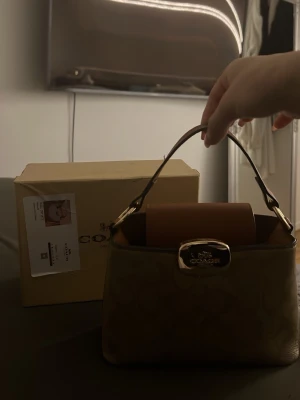 Beige handväska från Coach - Snygg beige handväska från Coach med klassiskt monogrammönster och guldfärgad logga på framsidan. Väskan har ett kort handtag i brunt läder och detaljer i guldmetall. Perfekt storlek för det viktigaste och ger en lyxig känsla till din outfit.