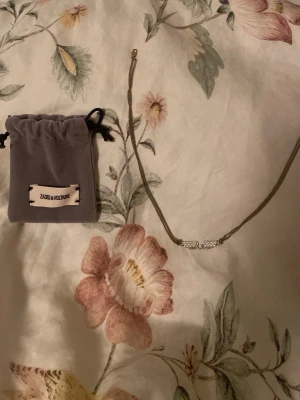 Zadig & Voltaire halsband - Snyggt halsband från Zadig & Voltaire i silverfärg med små vita pärlor på mitten av kedjan.  halsbandet kommer med en grå tygpåse med logga. Perfekt accessoar för att lyfta din outfit. Defekter: Kedjan är lite rostig som man ser på bild 2 men själva loggan är i fint skick 