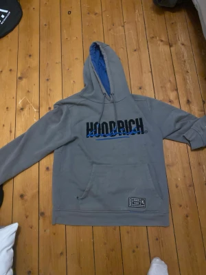 Grå hoodie från Hoodrich - Säljer en grå hoodie från Hoodrich med svart och blå logga på bröstet och tryck på ryggen. Hoodien har en stor känguruficka framtill, dragsko i huvan och blått innerfoder i huvan. Perfekt för en avslappnad streetwear-look.