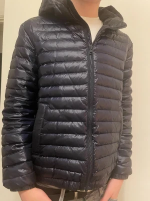 Marinblå lättviktsdunjacka med huva - Snygg svart dunjacka med quiltad design och huva. Jackan har dragkedja framtill, två sidofickor och en lätt, smidig passform. Perfekt för kyliga dagar och enkel att matcha med olika outfits.