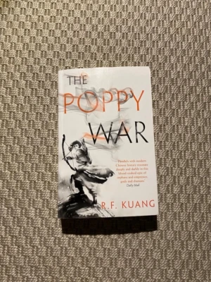 The Poppy War av R.F. Kuang - En engelskspråkig fantasyroman med vit och svart mjukpärm, dekorerad med illustrationer i gråskala och orange text. Boken utforskar teman som krig, magi och överlevnad i en fiktiv värld inspirerad av kinesisk historia.