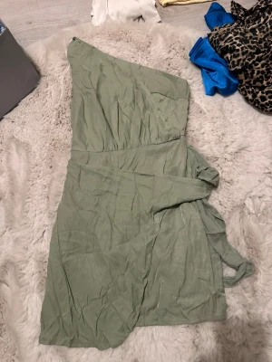 Grön oneshoulder klänning - Snygg ljusgrön oneshoulder klänning med omlottkänsla och knyt i midjan. Klänningen har en kort längd och är tillverkad i ett tunt, mjukt tyg som ger ett luftigt intryck. Perfekt för sommarens alla tillfällen.