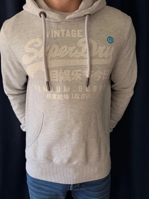 Ljusgrå hoodie från Superdry - Ljusgrå hoodie från Superdry med stor logga och japansk text på bröstet. Klassisk känguruficka framtill, ribbade muddar och justerbar huva med dragsko. Tillverkad i mjukt bomullsmaterial som känns skönt mot huden. Perfekt för en avslappnad och trendig look. Hör gärna av dig vid minsta funderingar.