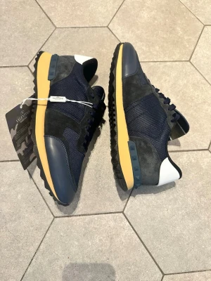 Marinblå sneakers från Valentino - Snygga marinblå sneakers från Valentino med ovandel i mesh och mocka, detaljer i läder och vit häl. Skorna har en gul sula med svarta partier och coola nitar på hälen. Perfekta för dig som vill ha en stilren men ändå unik look.