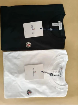 Moncler t-shirt svart och vit - Två stilrena t-shirts från Moncler, en i svart och en i vitt. Båda har rund hals, korta ärmar och Monclers ikoniska logga broderad på bröstet. Perfekta basplagg i mjuk bomull som passar till allt.