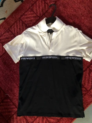 Svartvit skjorta från Emporio Armani - Snygg svartvit skjorta från Emporio Armani med tydlig logga längs midjan. Skjortan har klassisk krage, korta ärmar och är gjord i mjuk bomull. Perfekt för dig som gillar stilrena kontraster och exklusiva detaljer.passar till 12 år