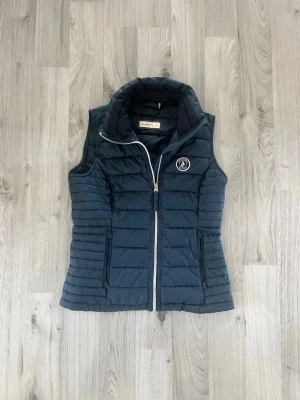 Mörkblå dunväst från abercrombie& fitch - Snygg mörkblå dunväst från abercrombie&fitch med vit dragkedja och två fickor med dragkedja. Västen har en hög krage och ett broderat märke på bröstet. Perfekt för lager på lager och ger en sportig look.