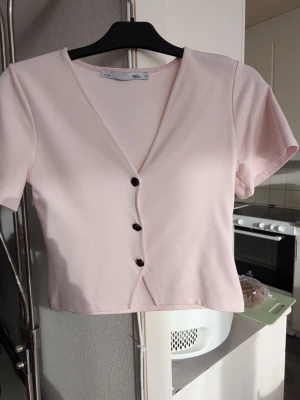 Ljusrosa topp XS  Lager 157 - Söt ljusrosa topp från Lager 157 med v-ringning och tre knappar framtill. Kortärmad och croppad modell i mjukt material, perfekt för en trendig och enkel look. Stlk XS 