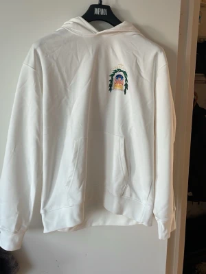 Vit hoodie med Casablanca-tryck - Säljer en vit hoodie från Avenida med ett färgglatt tryck på bröstet där det står 'CASABLANCA' och motiv av en byggnad omgiven av gröna blad. Hoodien har huva och stor magficka framtill. Perfekt för dig som gillar stilrena plagg med unik detalj.