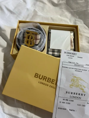 Burberry rutigt bälte med guldspänne - Snyggt Burberry-bälte med klassiskt rutigt mönster i beige, svart och rött. Bältet har ett stort guldigt 'TB'-spänne och är tillverkat i skinn. Perfekt accessoar för att lyfta din outfit med en lyxig touch. Plånbok med oxå 