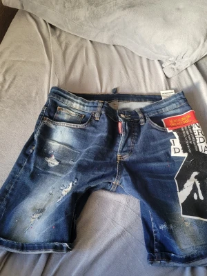 Dsquared byxor shorts  - Snygga jeansshorts i blå denim med trendig, sliten design. Shortsen har dekorativa slitningar och ett unikt tryck på ena benet som ger en modern och streetwear-inspirerad stil. Klassisk femficksmodell med bälteshällor och knappstängning. Perfekta för sommaren och enkla att matcha med t-shirt eller hoodie.