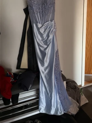 ljusblå balklänning  - Elegant ljusblå långklänning med vackra spetsdetaljer upptill och längs sidan. Klänningen har en figurnära passform upptill och faller snyggt ner mot golvet i ett glansigt material. Perfekt för speciella tillfällen när du vill känna dig extra fin.