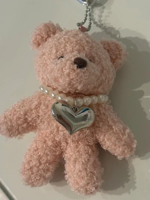 Nyckelring med rosa nalle och pärlhalsband - Söt nyckelring med en fluffig rosa nallebjörn i mjukt tyg. Nallen har ett pärlhalsband med ett stort silverfärgat hjärthänge. Perfekt att hänga på väskan eller nycklarna för en gullig touch.💘Aldrig använd! Orginalpris: 79kr