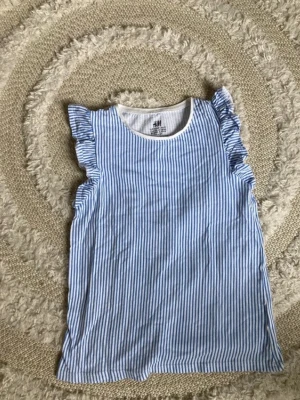 Blåvit randig linnetopp från H&M - Supersöt blåvit randig linnetopp från H&M med volangdetaljer vid axlarna. Toppen är ärmlös och har en rund halsringning. Perfekt till sommaren och enkel att matcha med jeans eller shorts. 💕