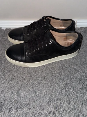 Lanvin sneakers i mocka och läder - Snygga svarta sneakers med ovandel i mocka och tåparti i slätt läder. Skorna har svarta skosnören, vita sulor och diskreta metalldetaljer vid snörningen. Insidan är ljusbeige och designen är klassiskt stilren. Skick 8/10 ända defekten är att dem är lite slitna innut i skon men verkligen inget som är sönder utan bara att foten har gjort avtryck (se bild). Hör av dig vid andra frågor, funderingar eller om du önskar fler bilder!😊 priset är diskuterbart!!
