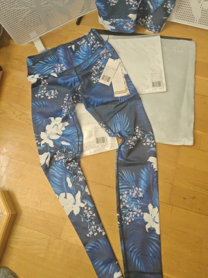 Blommigt träningsset från Stronger topp M Thights S Ny med lapp - Säljer ett snyggt träningsset från Stronger med blå botten och tropiskt blommönster i vitt och rosa. Setet består av en sport-bh och tights i stretchigt, snabbtorkande material. Perfekt för dig som vill träna med stil och sticka ut på gymmet!
