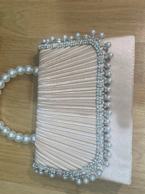 Ljusrosa kuvertväska med pärlhandtag - Snygg ljusrosa kuvertväska i satin med veckad framsida, dekorerad med glittrande strass och stora pärlor längs kanten. Handtaget består av stora vita pärlor som ger en lyxig känsla.