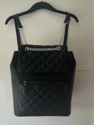 Som ny. Svart quiltad ryggsäck med kedjedetalj - Snygg svart ryggsäck i quiltat mönster med silverfärgad kedja på locket och dragkedjeförsedd ytterficka. Väskan har justerbara axelband och en stilren, modern look. Perfekt för dig som gillar trendiga accessoarer med lite edge. Använd en gång.
