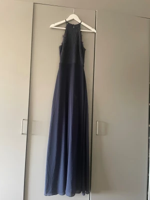 Marinblå långklänning med spets - Elegant marinblå långklänning med spetsdetaljer upptill och ärmlös design. Klänningen har en hög halsringning och ett flytande, långt kjolparti. Perfekt för speciella tillfällen där du vill känna dig extra fin.