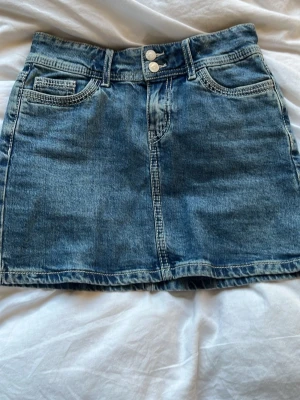 Blå jeanskjol med fickor - Snygg blå jeanskjol i klassisk modell med fem fickor, bälteshällor och dubbla knappar framtill. Kjolen har en rak passform och är tillverkad i slitstarkt denimtyg. Perfekt att matcha med en t-shirt eller hoodie.