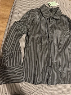 Randig svart skjorta med knytband - Snygg svart skjorta med vita ränder och knytband i midjan. Skjortan har långa ärmar, klassisk krage och knappar framtill. Tillverkad i ett mjukt bomullstyg som känns bekvämt och stilrent. Perfekt för dig som gillar en clean och modern look.