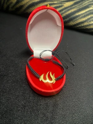 Svart armband med guldfärgad detalj - Snyggt armband med svart flätad tråd och en guldfärgad metallplatta med arabisk text. Justerbar knytning för perfekt passform. 