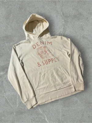 Beige hoodie från Denim & Supply | M - Snygg beige hoodie från Denim & Supply med tryck i rött framtill som visar texten 'DENIM & SUPPLY' och ett motiv av en person med fjäderprydd huvudbonad. Hoodien har magficka och ribbade muddar. Perfekt för en avslappnad streetwear-look.