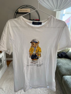 Vit Polo Bear t-shirt från Ralph Lauren - Vit t-shirt från Ralph Lauren med det ikoniska Polo Bear-motivet på bröstet. T-shirten har rund hals, korta ärmar och är gjord i mjuk bomull. Perfekt för dig som gillar klassisk streetstyle med en twist.