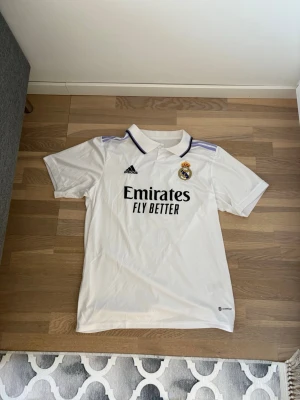 Real Madrid matchtröja Benzema 9 - Säljer en vit Real Madrid matchtröja från Adidas med svart tryck 'Emirates Fly Better' på bröstet och klubbmärke. På ryggen står det 'Benzema' och nummer 9. Tröjan har krage och lila detaljer på axlarna. Perfekt för fotbollsfans!