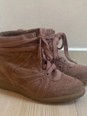 Rosa höga sneakers i mocka - Snygga höga sneakers isabel marant liknande i smutsig rosa mocka med perforerade detaljer och breda skosnören. Skorna har en diskret kilklack och gummisula som ger extra höjd och stil. Perfekta för dig som vill ha en trendig och bekväm look.