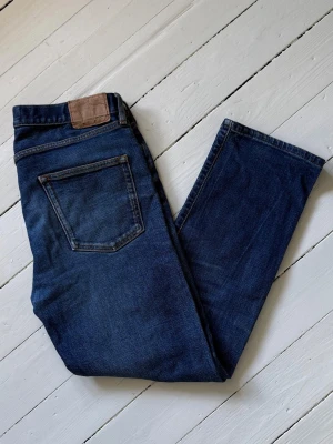 Jeanerica Straight Jeans - Straight jeans från Jeanerica. Säljer ett par klassiska mörkblå jeans med rak passform och femficksmodell. Jeansen har en snygg läderpatch bak i midjan och kontrastsömmar. Perfekta till en avslappnad stil och tillverkade i slitstark denim. Fråga om mått och storlek då dem har blivit skräddade.