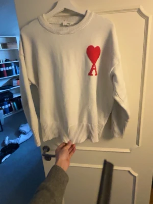 Vit stickad tröja Ami Paris hjärta - Vit stickad tröja från Ami Paris med röd hjärtlogga och bokstaven A på bröstet. Tröjan har rund halsringning, ribbade muddar och nederkant. Perfekt för dig som gillar stilrena plagg med en twist.
