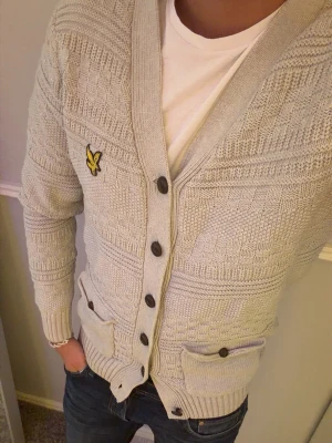 Ljusgrå kofta från Lyle & Scott - Snygg ljusgrå stickad kofta från Lyle & Scott med deras klassiska gula logga på bröstet. Koftan har v-ringning, fem knappar framtill och två fickor med knappdetaljer. Ribbstickade muddar och nederkant ger en schysst passform. Perfekt till jeans eller chinos. Cardigan