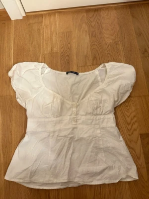 Brandy Melville blus - Söt vit blus med puffiga korta ärmar och dekorativa knappar framtill. Blusen har en djup rundad urringning och smock i ryggen för skön passform. Tillverkad i ett lätt tyg som ger en luftig känsla. Perfekt att styla med jeans eller kjol.