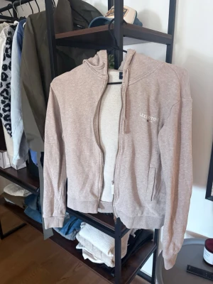 Beige hoodie från Lexington - Snygg beige hoodie från Lexington med dragkedja framtill och två fickor. Klassisk design med huva och diskret Lexington-logga på bröstet. Tillverkad i mjukt bomullsmaterial som känns skönt mot huden. Perfekt för en avslappnad och stilren look.