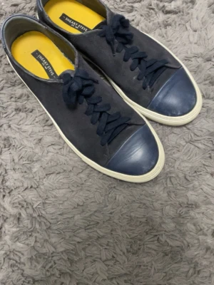 Mörkblå sneakers från Sneaky Steve - Snygga mörkblå sneakers från Sneaky Steve med ovandel i mocka och tåparti i gummi. Skorna har vita sulor och matchande mörkblå skosnören. Insidan är gul för en cool kontrast. Perfekta för dig som gillar stilrena och klassiska sneakers.