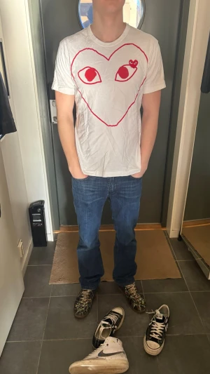 Vit Comme des Garçons Play t-shirt - Vit t-shirt från Comme des Garçons Play med det ikoniska röda hjärtat och ögon-trycket på bröstet. Klassisk passform och rund halsringning. Perfekt statement-plagg för dig som gillar streetwear och vill sticka ut.