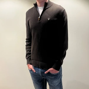 Brun halvzip-tröja från Morris - Snygg brun stickad tröja från Morris med halv dragkedja och ribbade muddar. Tröjan har en liten broderad logga på bröstet och hög krage med knappdetalj. Perfekt för en stilren och avslappnad look.