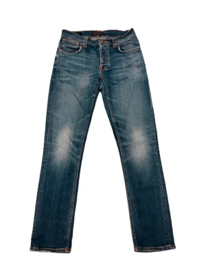 Blå raka jeans från Nudie Jeans - Säljer ett par klassiska blå jeans från Nudie Jeans med raka ben och snygg slitning på framsidan. Jeansen har fem fickor, orangea sömmar och detaljerade bakfickor med Nudie-loggan. Tillverkade i bomull för en skön och avslappnad look.