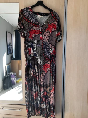 Mönstrad långklänning med kort ärm från Odd Molly - Unik långklänning med färgstarkt blommigt och geometriskt mönster i svart, rött och beige. Klänningen har v-ringning, knappar framtill och korta ärmar. Perfekt för dig som vill sticka ut med en bohemisk och cool stil.