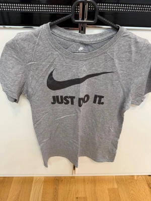 Grå Nike t-shirt med tryck - Snygg grå t-shirt från Nike med det klassiska swoosh-loggan och texten 'Just Do It.' på bröstet. T-shirten har rund halsringning och korta ärmar. Tillverkad i mjuk bomull som känns skön mot huden.