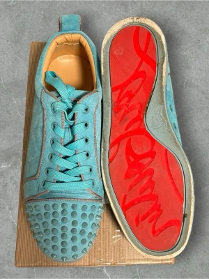Blå Louboutin sneakers med nitar - Unika turkosa sneakers från Christian Louboutin i mocka med coola nitar på tån och ikonisk röd sula. Skorna har turkosa skosnören och beige innersula. Perfekta för dig som vill sticka ut med en edgy och lyxig stil. En bit borta ingen box